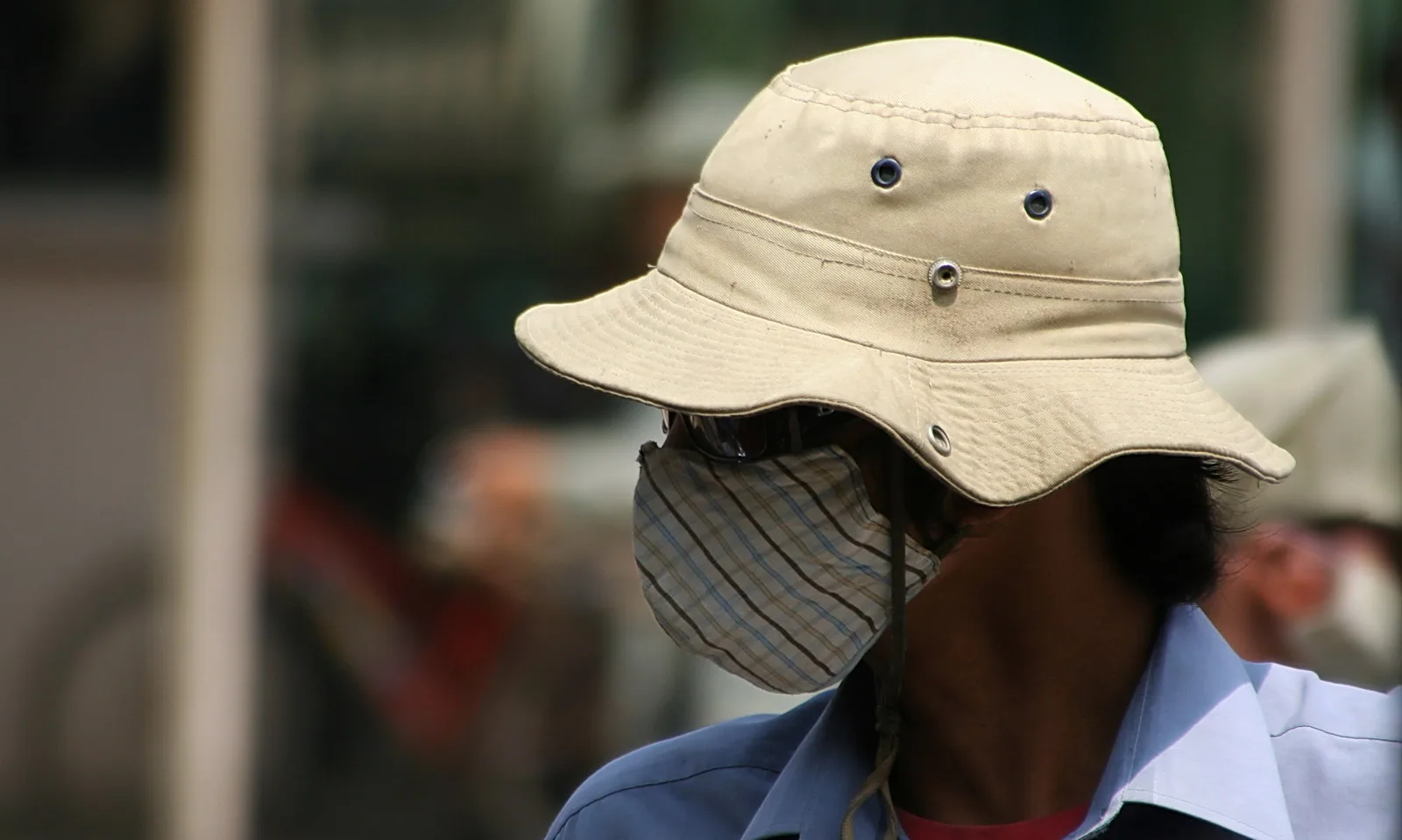 Mann mit beigem Buschhut und gestreifter Stoffmaske vor unscharfem Straßenhintergrund, im Profil nach links gewandt.