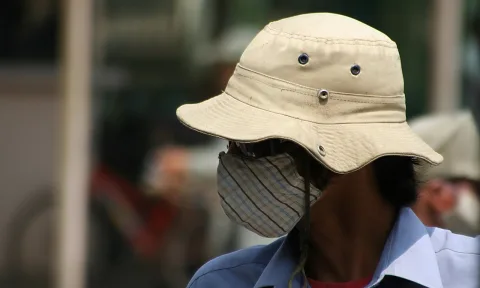 Mann mit beigem Buschhut und gestreifter Stoffmaske vor unscharfem Straßenhintergrund, im Profil nach links gewandt.