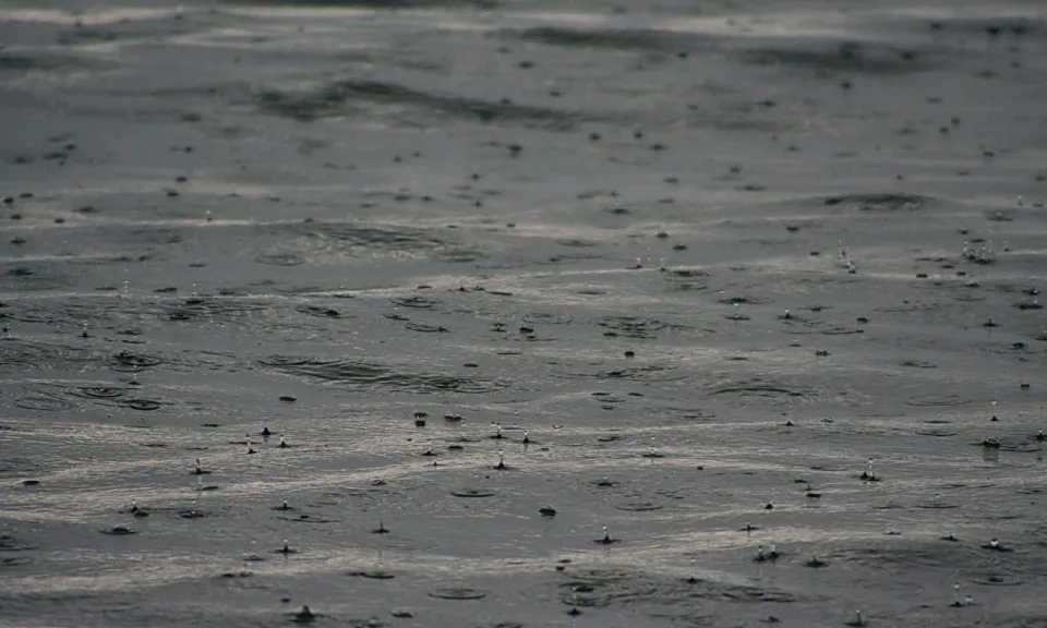 Regen fällt auf eine graue Wasseroberfläche, überall entstehen kleine Spritzer und Kreise.