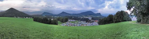 Panoramablick über eine grüne Wiese auf einen Zeltplatz im Tal, dahinter bewaldete Berge unter bewölktem Abendhimmel.