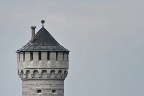 Oberer Teil eines runden Steinturms mit spitzem Schieferdach, Schornstein und Kugelspitze vor grauem Himmel.
