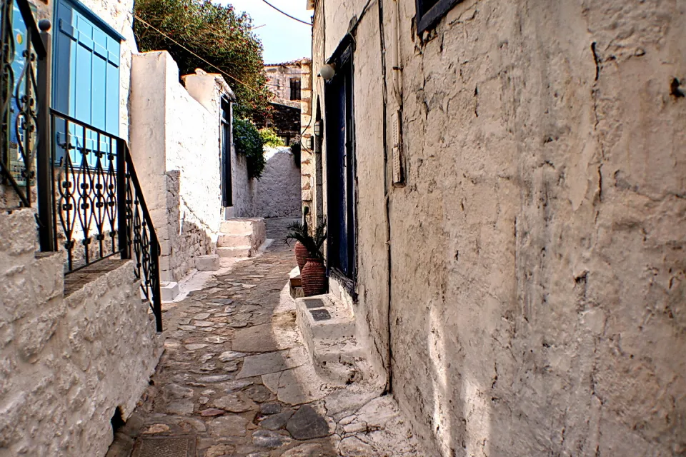 Schmale Gasse mit Kopfsteinpflaster zwischen weiß getünchten Mauern, links ein blauer Fensterladen und ein schmiedeeisernes Geländer, rechts zwei Tontöpfe vor einer dunklen Tür.