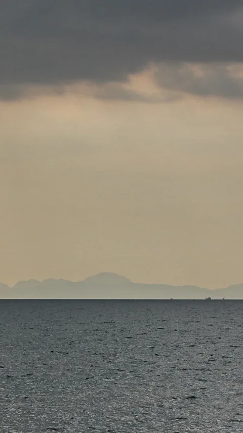 Dunkle Wolkenbank über einem Meer, am Horizont zeichnen sich blaugraue Berge und zwei winzige Schiffe ab, der Himmel ist beige-grau getönt.