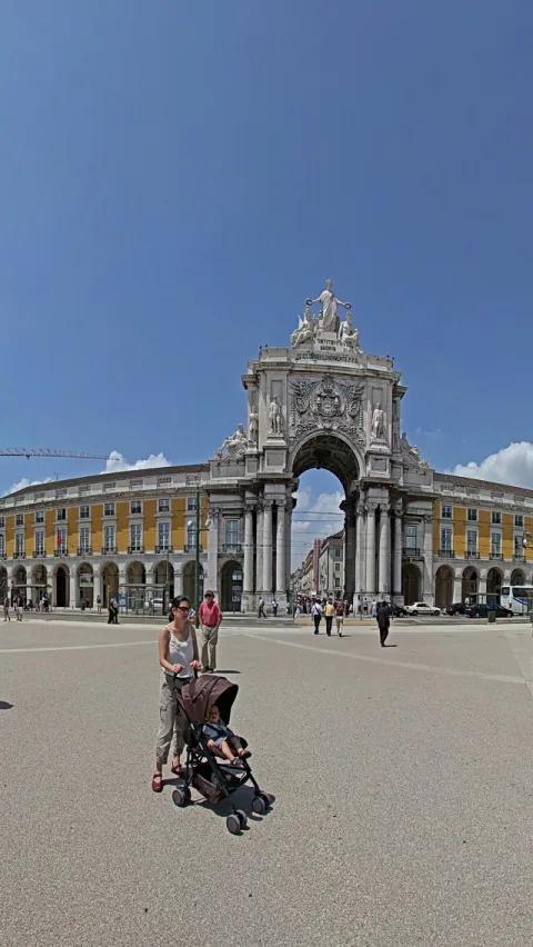 Großer weißer Triumphbogen mit Skulpturen auf einem weiten Platz in Lissabon, davor eine Frau mit Kinderwagen, dahinter Passanten und gelb gestrichene Arkadenhäuser unter blauem Himmel.