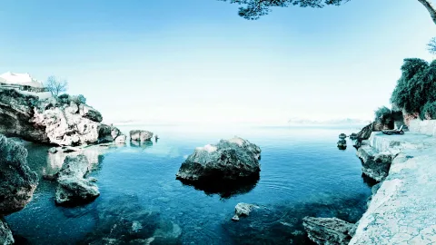 Ruhige türkisblaue Meeresbucht mit großen Felsen im Wasser, links ein weißes Zeltdach, rechts ein Liegestuhl auf einer Steinterrasse und ein Baum im oberen Bildrand.