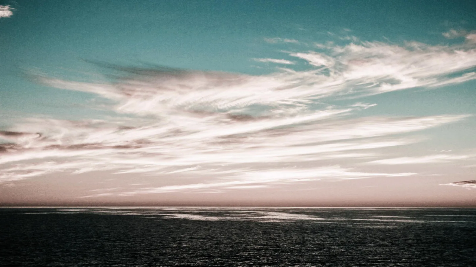 Schmale, helle Federwolken ziehen über einen türkisfarbenen Himmel, darunter liegt eine dunkle, ruhige Meeresfläche unter einem rosafarbenen Horizont.