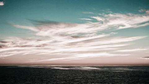 Schmale, helle Federwolken ziehen über einen türkisfarbenen Himmel, darunter liegt eine dunkle, ruhige Meeresfläche unter einem rosafarbenen Horizont.
