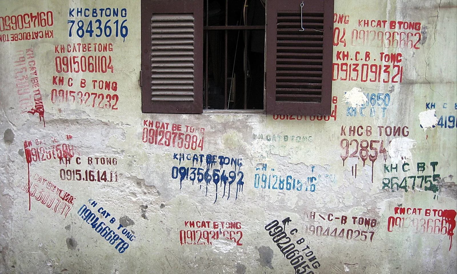 Verwitterte Hauswand mit aufgemalten Telefonnummern und Schriftzügen in Rot, Blau und Grün, oben ein Fenster mit braunen Holzläden.