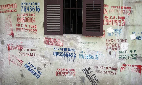 Verwitterte Hauswand mit aufgemalten Telefonnummern und Schriftzügen in Rot, Blau und Grün, oben ein Fenster mit braunen Holzläden.