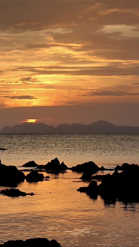 Sonnenuntergang über dunklen Felsen im Meer, dahinter eine Bergkette am Horizont.